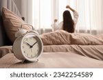 It's lazy morning o'clock. Alarm clock on bedside table and woman stretching in room, selective focus
