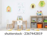 Small photo of Table with chairs, shelves and toys near white wall in playroom. Stylish kindergarten interior