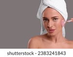 Young woman massaging her face with rose quartz roller on grey background, space for text
