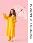 Small photo of Young African-American woman wearing yellow raincoat and gumboots with umbrella on pink background