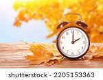 Small photo of Alarm clock and autumn leaves on table outdoors. Daylight saving time end