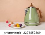 Electric kettle and plate with macaroons on table near brown wall