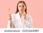 Small photo of Young woman holding spatula with sugaring paste and razor on pink background
