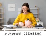 Small photo of Young female student calculating finances at table with piggy banks. Student loan concept