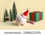 Piggy bank in festive party hat with Christmas trees, gift box and candy canes on beige background