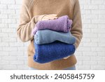 Woman holding stack of different stylish sweaters on light brick background