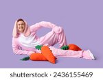 Happy young man in Easter bunny costume lying with plush carrots on lilac background