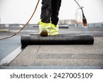 Small photo of professional roofer applying tar with blowtorch on new roofing felt for waterproofing a flat roof construction site with safety gear