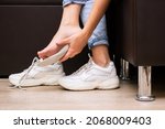 Pain in the feet from uncomfortable shoes. A woman i a white sneakers tries on a shoe insole sitting on a couch. Discomfort and ache caused by walking for a long time. Orthodontics