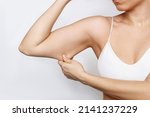 Small photo of Close-up of a young caucasian woman grabbing skin on her upper arm with excess fat isolated on a white background. Pinching the loose and saggy muscles. Overweight, liposuction concept