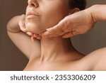 Small photo of Cropped shot of young caucasian woman touching under the chin with hands massaging her face on dark brown background. Rejuvenation, facelift, facefitness. Exercises from the second chin, pelican neck