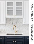 Small photo of A kitchen sink detail shot in a white and blue kitchen with a gold faucet, marble countertop, and small marble subway tile backsplash.