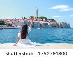 Young tourist woman sitting on Harbour holding her hand up wave to beautiful rovinj city, Croatia. Happy traveler girl enjoy on vacation in Europe. Freedom lady with white dress on a sunny day.