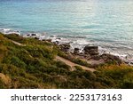 Rocky sea coast with sea waves and green bushes on the slopes. Sand on the shore, rocky boulders in the sea. The natural landscape of the island of Rhodes