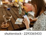 Beautiful young woman is guessing on cards with tarot, runes on wooden table and uses an online app in phone to interpret divination, home interior,candles and dry herbs..