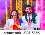 Happy young couple with color on face saying namaste againt flower decorated background - concept of holi festival greeting, wishes and Indian traditional culture
