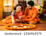 Small photo of Happy indian husband helping to wife decorating rangoli with flowers for diwali festival celebration while sitting on floor at home - concept of relationship, bonding, and religious ceremony