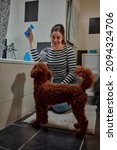 Small photo of Woman sitting on the floor in the bathroom and trying to seduce puppy with toy to get in bathtub
