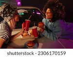 Small photo of Two joyful retro stylish girls are outdoors eating takeout food on the hood of their car hood.