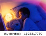 Little preschool kid boy sleeping in bed with colorful night led lamp. School child dreaming and holding plush toy. Kid angry of darkness.