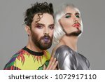 Small photo of A transgender couple, 2 attractive male persons with beards and makeup on their faces, looking at the camera. The guy with a quiff touching the back of his partner in women's clothing and bob-cut wig.