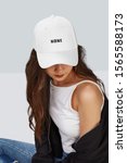 Small photo of Cropped upward shot of a dark-haired girl, wearing white baseball cap with lettering "none", white tank top, jeans with scuffs and black jacket. The cap peak is closing upper part of a face.