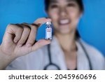 Small photo of Female doctor wearing a white coat with a stethoscope around her shoulder holding a glass vial of PCV or Pneumococcal conjugate vaccine, smiling. Blue background. Healthcare And Medical concept.