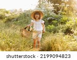 Small photo of Portrait of smiling little girl walks in the rays of a sunset in a flowering meadow, enjoying the summer, warmth, flowers, freedom. Child with straw hat and bag is having fun outdoors.