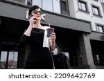 Successful businesswoman leader stands in big city talks on mobile phone. Smiling woman making business call on cell walking with cup of coffee on background of office centre, multitasking outdoors.