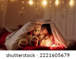 Small photo of little cute smiling baby girl with white teddy bear peeking out from under a blanket on a bed at home with lighting garlands at dark. Child looking in camera. Fairytale childhood with miracle concept.