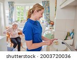 Small photo of Female Home Help Cleaning House Doing Washing Up In Kitchen Whilst Chatting With Senior Woman