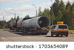 Small photo of Long oversized cargo transportation cogistics, big large pipes on 5-axle lowbed trawls semi-trucks with an escort car on highway at summer day