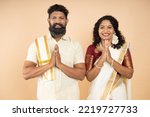 Happy South indian couple wearing traditional white dress standing in a greeting pose to Namaste hands isolated on beige background, man wearing lungi and woman wearing saree.