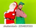 Photo of fat bearded santa claus crossing his arms on green chroma background next to his elf friend. Santa claus and elf with arms crossed