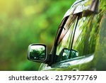 Rear view of a silver SUV car driving on a forest road, green trees reflected on the mirror, green forest blurred in the background. Road trip, holidays concepts. Focus on the side mirror.