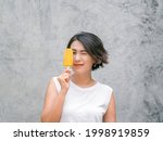 Woman eating popsicles. Happy beautiful Asian woman short hair wearing casual white sleeveless shirt holding yellow popsicle, outdoors. Smiling female enjoying ice lolly in summer.