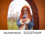 Young woman with bright hairstyle in warm clothes browsing mobile phone, standing leaning on iron installation in park in snowy weather