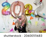 young woman celebrating new year 2022 at home quarantine. smiling girl holding cute british cat in shirt. big number balloons. silvester party. december 31. lockdown. confetti falling.  january 1