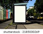bus shelter blank ad panel. billboard display. empty white lightbox sign at busstop. glass and aluminum structure. city transit station. urban street and green park setting. outdoor advertising.