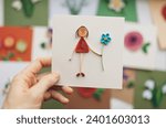 Small photo of Hand holding quilling card with stick figure girl giving flower. woman making greeting cards. Hand made of paper quilling technique. Handicraft at home. Hobby, home office.