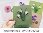 Small photo of Hand holding quilling card with Snowdrops flowers. woman making greeting cards. Hand made of paper quilling technique. Handicraft at home. Hobby, home office.