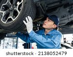 Asian automotive mechanic repairman look under car condition in garage. Vehicle service guy worker doing check or mend car wheel and work in mechanics workshop with confidence to repair car engine car