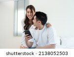 Small photo of Asian new marriage couple sit on bed and look at telephone in bedroom. Attractive beautiful young man and woman in pajamas enjoy early morning activity and holding phone and gossip together at home.
