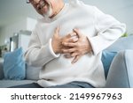 Small photo of Asian senior older man having chest pain feel suffer from heart attack. Attractive older mature patient has difficulty breath clutching his chest from acute pain while sitting on sofa in living room.