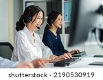 Two Asian beautiful businesswoman call center work in office corporate. Attractive young employee worker people sit on table, wear headset and use laptop computer talk to support customer at workplace