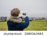 Young man in blue coat is taking a picture of city and green field by his phone. Cloudy day in Norway.