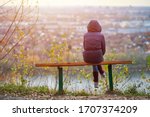 Small photo of Young woman sitting on bench in autumn city park and looking at cityscape, back view. Self-reflection of girl, thinking about unrequited love for Valentines Day. Introspection concept.