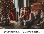 Young teen boy with long hair thoughtful look sad eyes negative mood angry and crying at home. Stylish zoomer gen Z pensive on new year holidays with xmas tree bokeh lights garlands eve 25 december