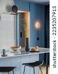 Small photo of Interior design of kitchen interior with marble kitchen island, blue wall, black chokers, bowl with fruits, big window, cup, wooden floor, lamp on wall and personal accessories. Home decor. Template.