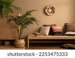 Small photo of Warm and cozy ethno living room interior with couch, patterned pillows, plants i flowerpots, fern, rattan sideboard, basket on wall, wooden bowl and personal accessories. Home decor. Template.